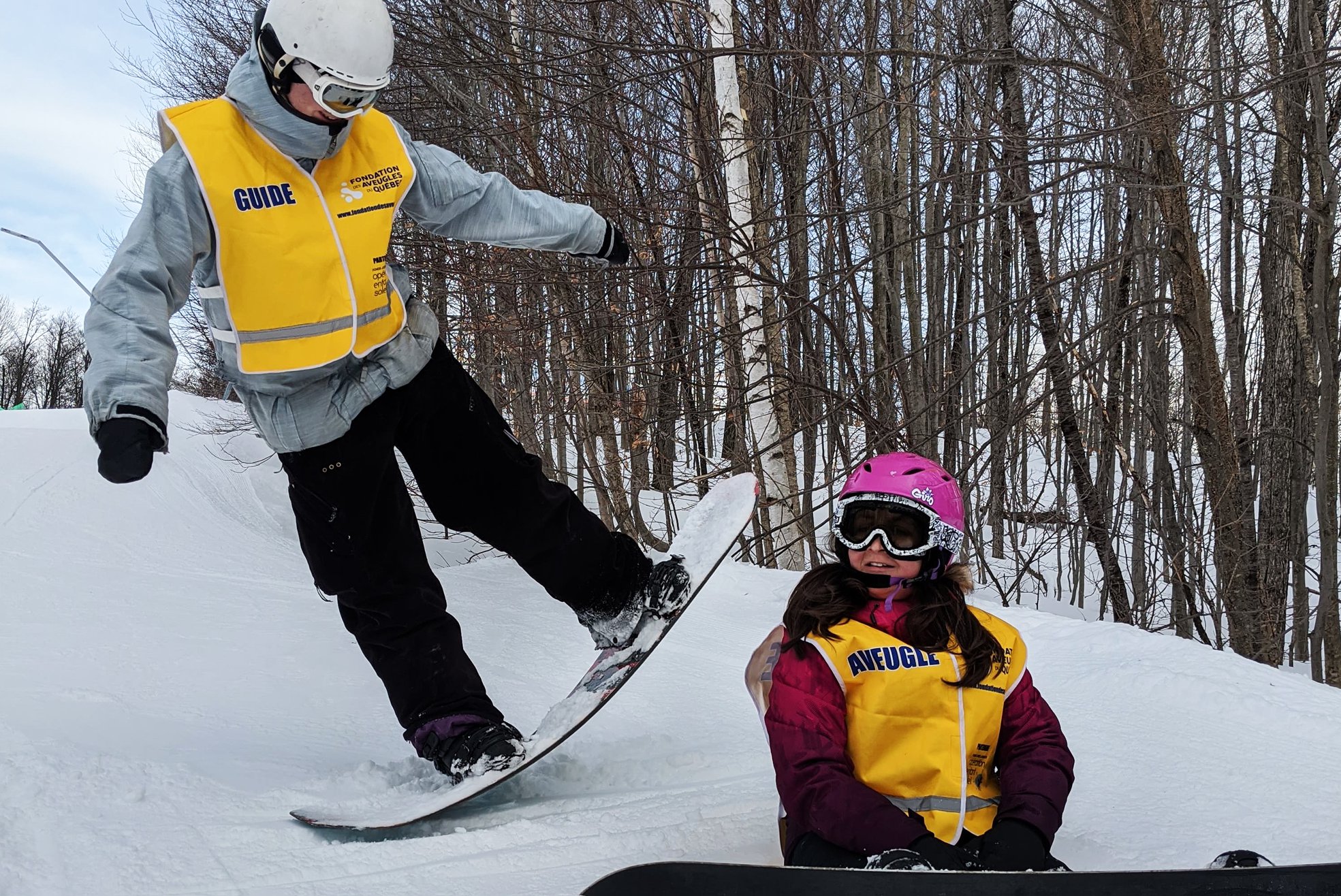 Un accompagnateur soulève un côté de sa planche à neige alors qu’une participante est assise dans la neige