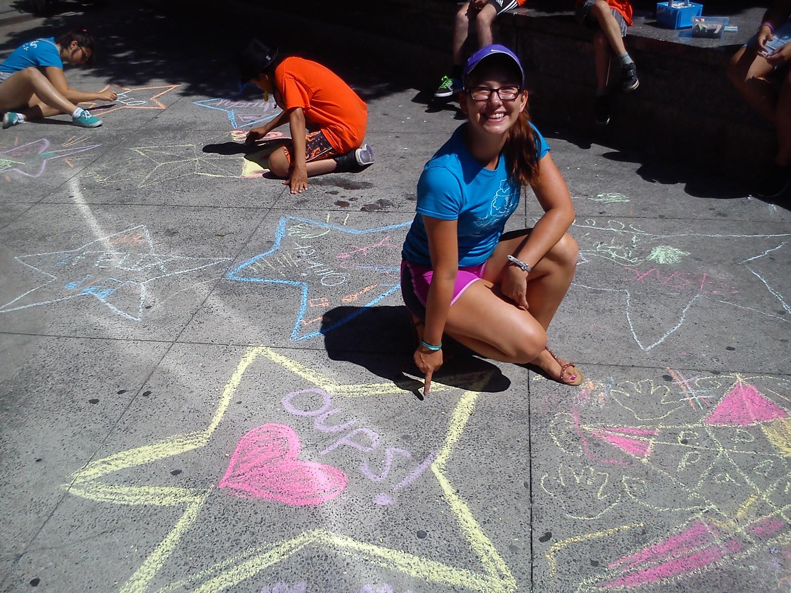Jessica est accroupie devant un dessin sur l’asphalte, à la craie, d’une étoile avec un cœur et le texte « Oups! » au centre.