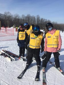 Une accompagnatrice et un participant se tiennent en accolade sur la piste de ski alors qu’un accompagnateur et une participante se tiennent derrière eux.