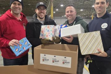 Quatre hommes souriants, vêtus de vêtements d'hiver décontractés, déposent des cadeaux de Noël emballés dans un grand carton portant des autocollants "Paniers de Noël