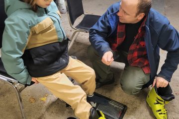 Un adulte guide aide un enfant à essayer des bottes de ski dans un espace intérieur.